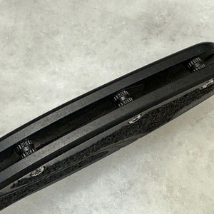 May include: Close-up of the black handle of a folding knife. The handle has a textured surface and a black metal clip.