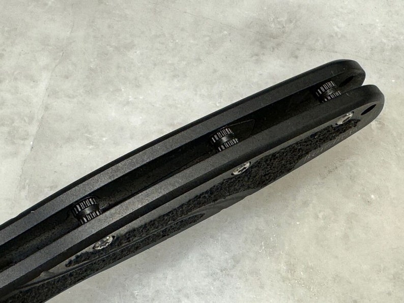 May include: Close-up of the black handle of a folding knife. The handle has a textured surface and a black metal clip.