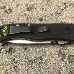 May include: A black folding knife with a green pocket clip and green screws. The knife has a black handle and a silver blade.