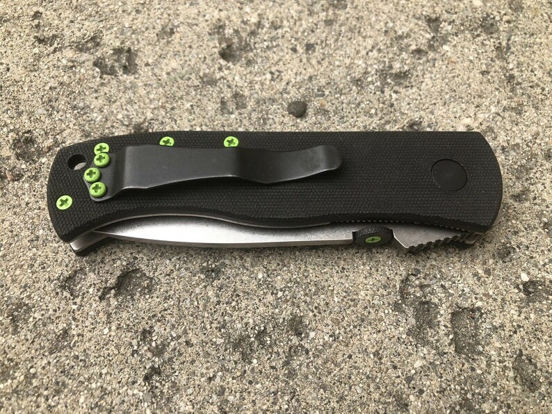 May include: A black folding knife with a green pocket clip and green screws. The knife has a black handle and a silver blade.