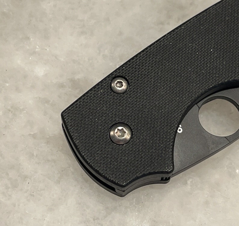 K&ouml;nnte beinhalten: Nahaufnahme eines schwarzen Taschenmessers mit zwei silbernen Schrauben. Das Messer hat einen strukturierten schwarzen Griff und eine silberne Klinge.