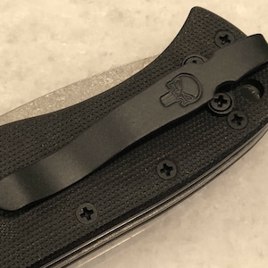 May include: Black pocket knife with a black pocket clip and a skull logo. The knife has a textured black handle and multiple screws.