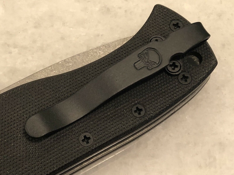 May include: Black pocket knife with a black pocket clip and a skull logo. The knife has a textured black handle and multiple screws.