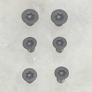May include: A set of six black metal screws arranged in two columns of three. Each screw features a flat, circular head with a six-pointed star-shaped torx drive recess in the center, designed for secure fastening applications in various projects.