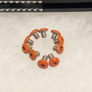 Puede incluir: Un conjunto de tornillos con cabezas naranjas dispuestos en círculo. Los tornillos tienen una hendidura en forma de estrella en la cabeza y un vástago roscado plateado. Fondo claro y neutro.