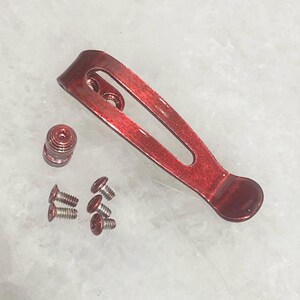 Pu&ograve; includere: Un set di componenti per clip da tasca metalliche rosse, tra cui una clip, una vite e diverse viti piccole. La clip ha un design curvo con un'estremit&agrave; arrotondata. I componenti sono disposti su una superficie chiara.