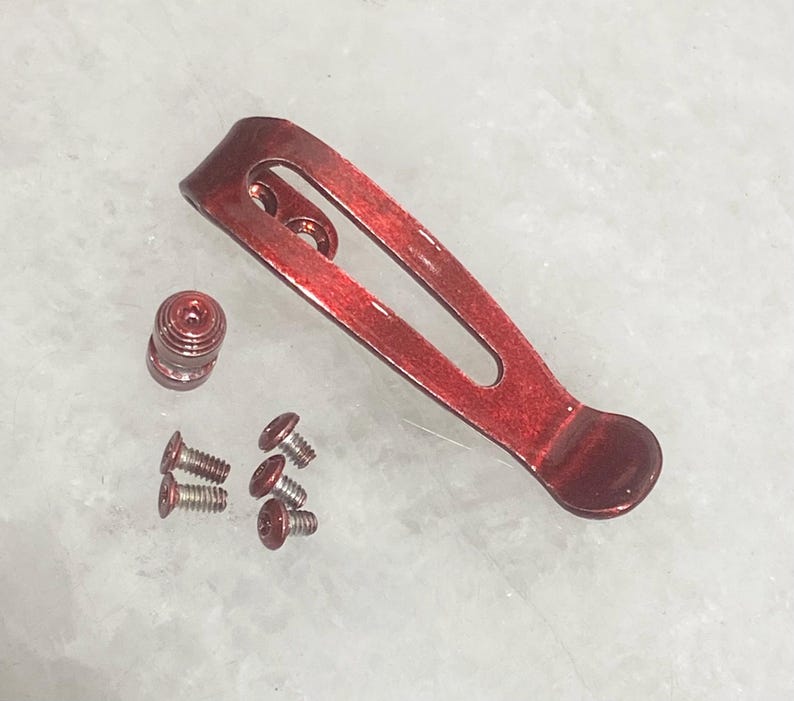 Pu&ograve; includere: Un set di componenti per clip da tasca metalliche rosse, tra cui una clip, una vite e diverse viti piccole. La clip ha un design curvo con un'estremit&agrave; arrotondata. I componenti sono disposti su una superficie chiara.