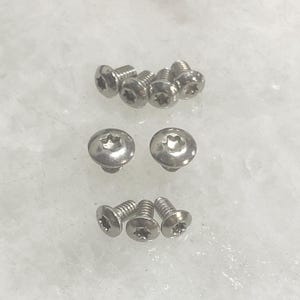 May include: A collection of eight silver-colored metal screws with rounded heads and star-shaped Torx drive recesses. The screws are arranged in three distinct rows, showcasing their threaded shanks and polished metallic finish in a clear view.