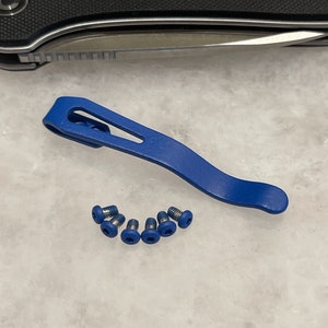 May include: A blue metal pocket clip with a curved design and a set of five small blue screws with star-shaped drive heads. The clip features an elongated cutout slot and a smooth finish, accompanied by matching hardware for secure attachment.