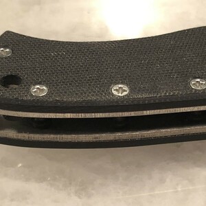 May include: Close-up of a black knife handle with a textured surface and silver accents. The handle has several small screws.