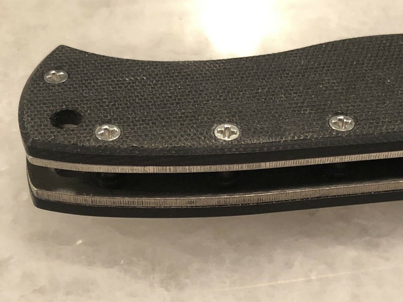 May include: Close-up of a black knife handle with a textured surface and silver accents. The handle has several small screws.