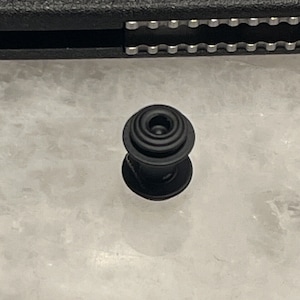 May include: A black plastic snap fastener with a circular design. The fastener has a raised center and multiple concentric rings.