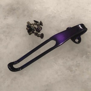 Peut inclure: Un clip de poche en métal violet pour un couteau ou un multi-outil. Le clip est fait d'un métal durable et a une finition lisse. Il est conçu pour être attaché à une ceinture ou à une poche.