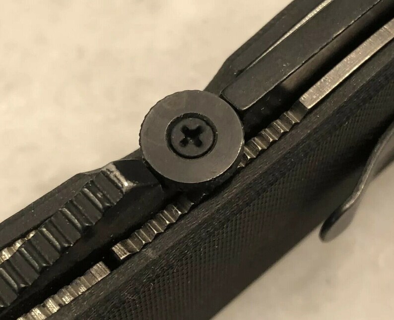 May include: Close-up of a black pocket knife with a textured handle and a silver blade. A black screw with a cross-shaped head is visible on the handle.