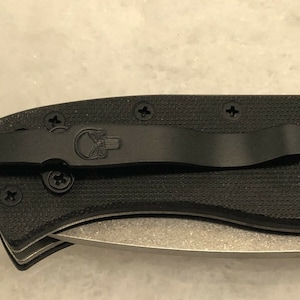 May include: A black folding knife with a black pocket clip and a skull logo. The knife has a textured black handle and a silver blade.