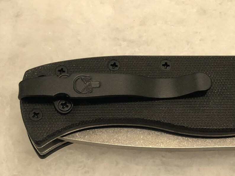 May include: A black folding knife with a black pocket clip and a skull logo. The knife has a textured black handle and a silver blade.