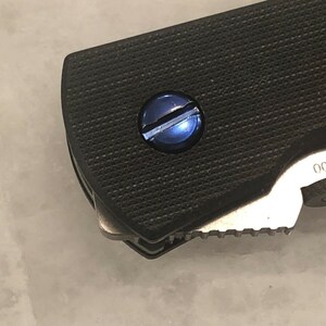 May include: Close-up of a black folding knife with a textured handle and a blue screw. The blade is partially visible, showing a serrated edge. The number "3300" is printed on the knife.