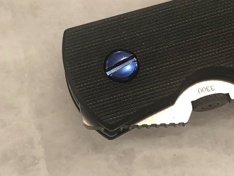 May include: Close-up of a black folding knife with a textured handle and a blue screw. The blade is partially visible, showing a serrated edge. The number "3300" is printed on the knife.