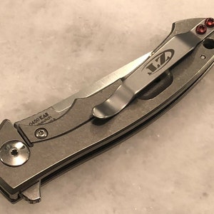 May include: A silver pocket knife with a black handle and a clip on the side. The clip has the letters "MZ" engraved on it. The knife has a small red screw on the side.