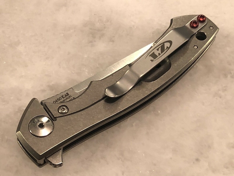 May include: A silver pocket knife with a black handle and a clip on the side. The clip has the letters "MZ" engraved on it. The knife has a small red screw on the side.