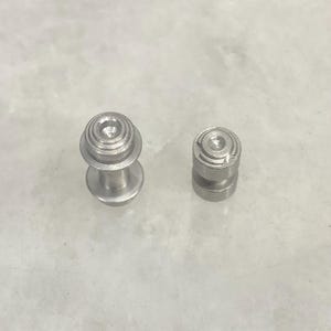 May include: Two small, silver-colored metal objects. One is taller with a rounded top and a stem, the other is shorter and cylindrical. Both have a series of concentric circles on their tops.
