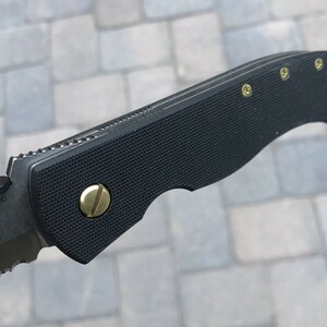 May include: A black folding knife with a serrated blade and a black handle with gold screws. The handle has a textured surface and the blade has the text "3300" on it.