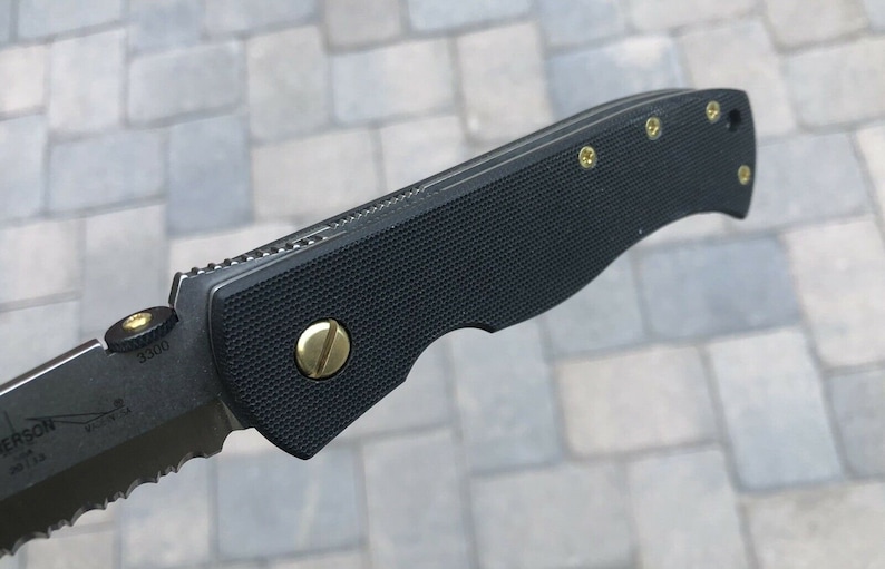 May include: A black folding knife with a serrated blade and a black handle with gold screws. The handle has a textured surface and the blade has the text "3300" on it.