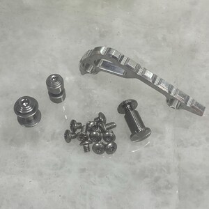 May include: An assortment of silver-colored metal hardware components, including screws, cylindrical pieces, and a slotted, elongated piece. The items are arranged on a light-colored surface, suggesting a mechanical or industrial theme.