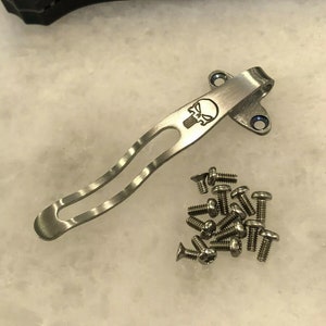 May include: A silver pocket clip with a skull design and a set of screws.