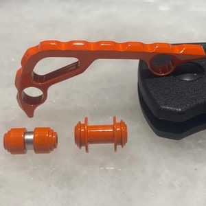 Puede incluir: Herramienta de metal naranja con un mango curvo y dos piezas de metal naranja más pequeñas. La herramienta tiene un agujero en el mango y las piezas más pequeñas tienen una pieza de metal plateada en el medio.