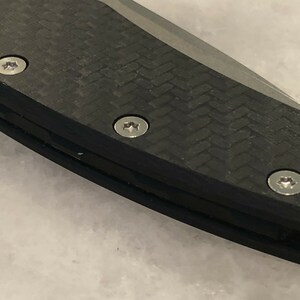 以下が含まれることがあります： 黒いカーボンファイバー製のナイフのハンドルを3本の銀色のネジで留めたクローズアップ。