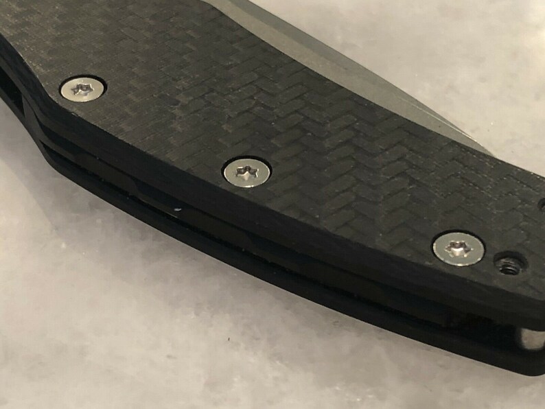 以下が含まれることがあります： 黒いカーボンファイバー製のナイフのハンドルを3本の銀色のネジで留めたクローズアップ。