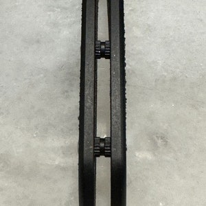 May include: Close-up of a black metal object with three sets of small black gears. The object is on a white surface.