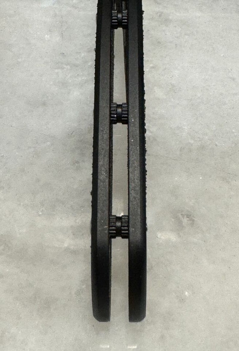May include: Close-up of a black metal object with three sets of small black gears. The object is on a white surface.