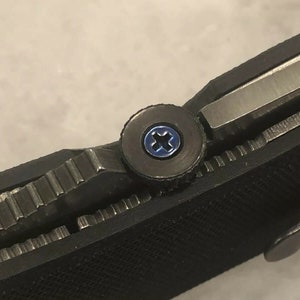 May include: Close-up of a black pocket knife with a blue screw. The screw is a Phillips head screw.
