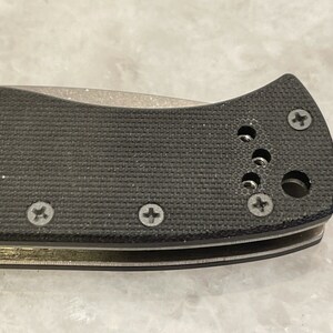 May include: Close-up of a black knife handle with a textured surface and multiple screws. The handle has a rounded shape and a small hole near the top.