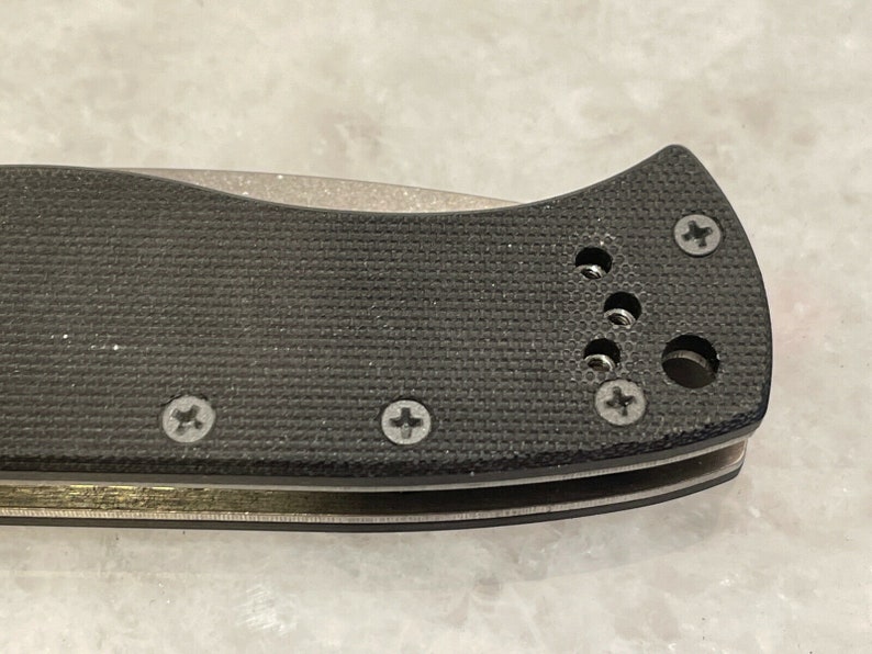 May include: Close-up of a black knife handle with a textured surface and multiple screws. The handle has a rounded shape and a small hole near the top.