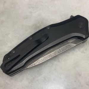 May include: A black folding knife with a partially serrated blade. The knife has a black handle with a textured surface and a pocket clip.