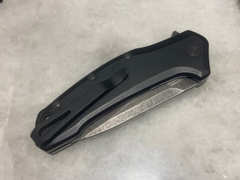 May include: A black folding knife with a partially serrated blade. The knife has a black handle with a textured surface and a pocket clip.
