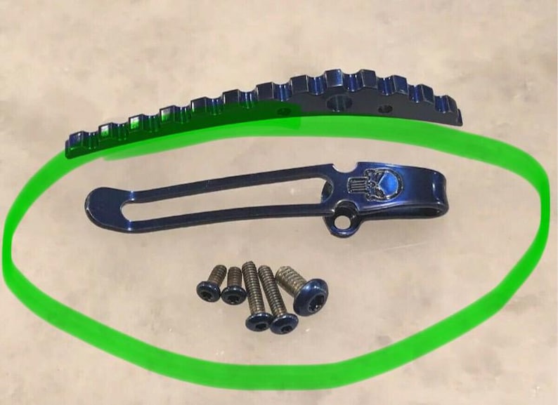 May include: A blue metal pocket clip with a skull design. The clip is on a green background with five small screws.