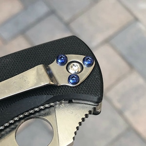 May include: Close-up of a black pocket knife with a silver clip and blue screws. The knife has a textured black handle and a silver blade.