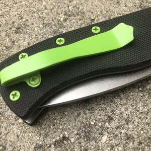 Pode incluir: Uma faca dobr&aacute;vel preta com um clip de bolso verde e uma l&acirc;mina prateada. A faca tem uma al&ccedil;a preta texturizada e cinco parafusos.