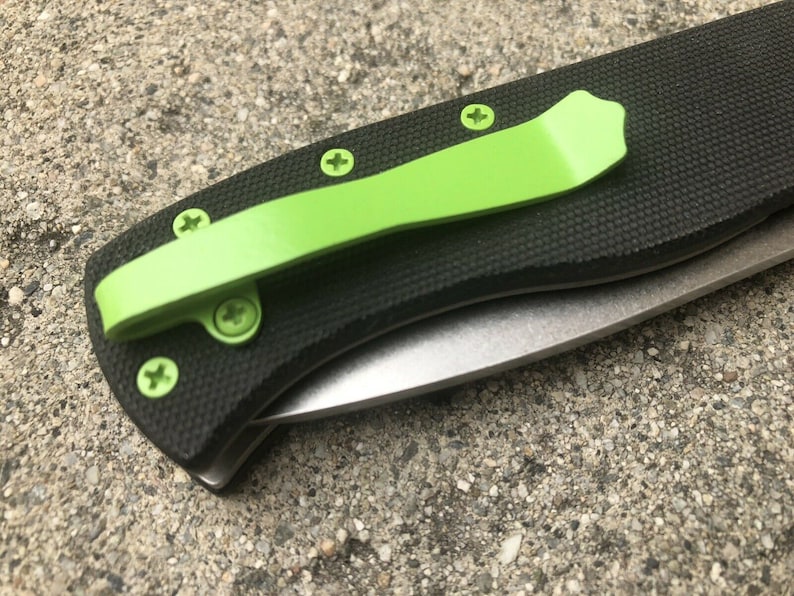 Pode incluir: Uma faca dobr&aacute;vel preta com um clip de bolso verde e uma l&acirc;mina prateada. A faca tem uma al&ccedil;a preta texturizada e cinco parafusos.
