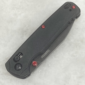 Pu&ograve; includere: Un coltello pieghevole nero con dettagli rossi. Il coltello presenta un manico strutturato, una lama e viti rosse. Il marchio "Kershaw" &egrave; visibile sulla lama. Il coltello &egrave; chiuso e pronto all'uso.