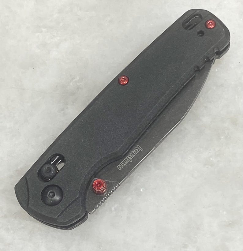 Pu&ograve; includere: Un coltello pieghevole nero con dettagli rossi. Il coltello presenta un manico strutturato, una lama e viti rosse. Il marchio "Kershaw" &egrave; visibile sulla lama. Il coltello &egrave; chiuso e pronto all'uso.