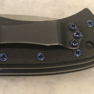 May include: Close-up of a black pocket knife with blue screws. The knife has a clip and a textured handle.