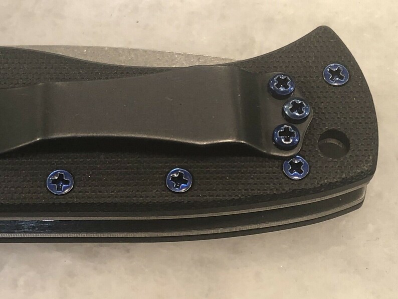 May include: Close-up of a black pocket knife with blue screws. The knife has a clip and a textured handle.