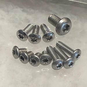 Puede incluir: Una colección de tornillos plateados con cabeza de estrella. Los tornillos están dispuestos en un patrón aleatorio sobre una superficie blanca.