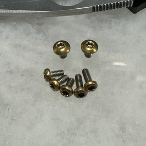 May include: A set of seven gold-colored screws with a star-shaped head. The screws are arranged in a semi-circle on a white surface.