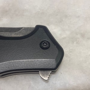 May include: Close-up of a black pocket knife with a textured handle and a silver blade. A small screw with a star-shaped head is visible on the handle.